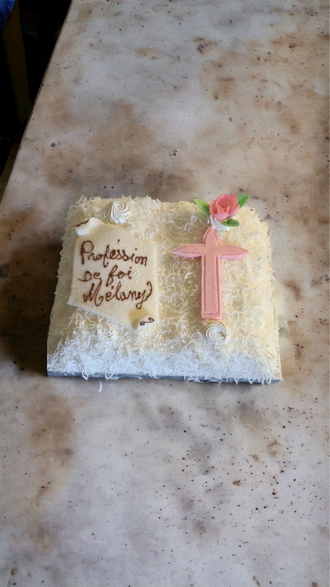 Gâteau blanc carré, enrobé de noix de coco, avec une croix rose et un parchemin avec du texte.