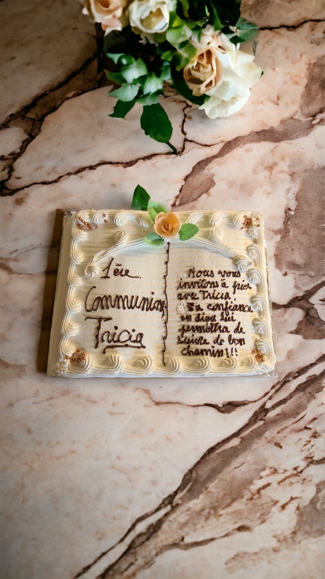 Gâteau décoré de fleurs et portant l'inscription « 1a Communione Tricio ». Couleurs : blanc, or et marron.