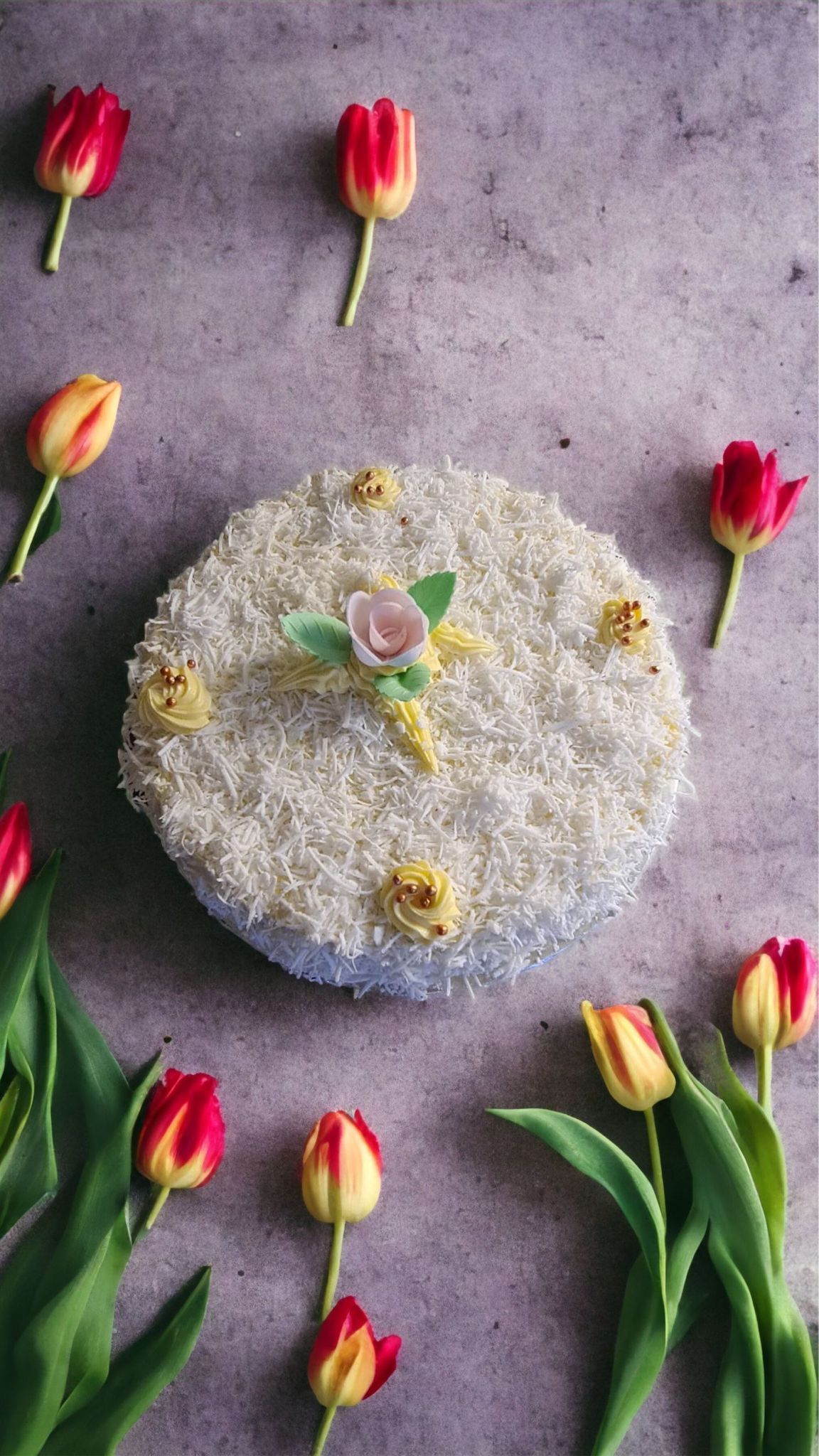 Gâteau avec glaçage blanc, entouré de tulipes rouges et jaunes sur une surface grise.