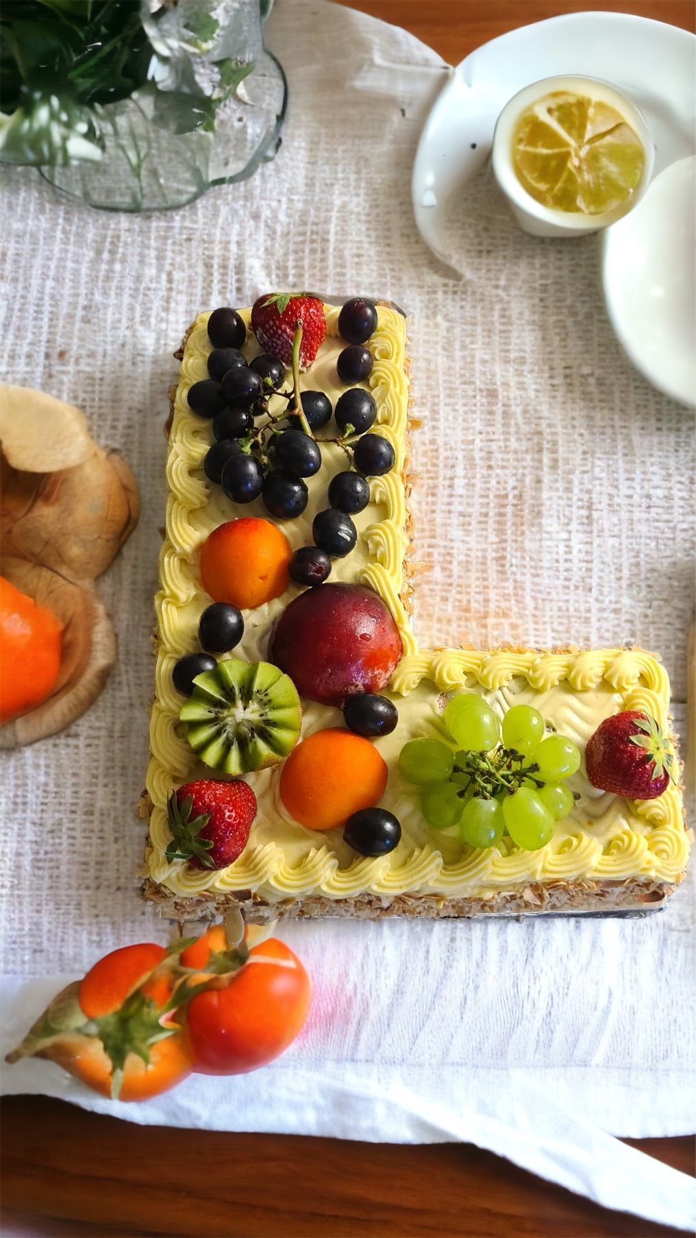 Gâteau lettre « L » décoré de fruits ; oranges, baies, raisins sur une nappe blanche.