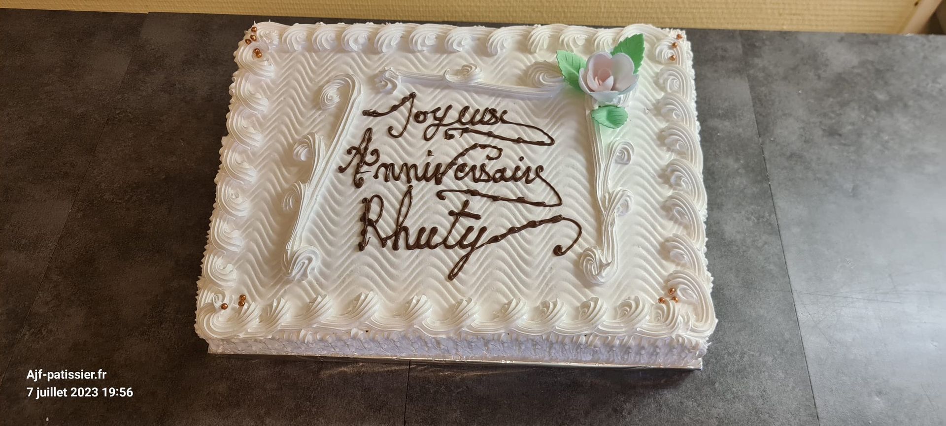 Un gâteau glacé blanc avec les mots «Joyeux Anniversaire Rhuty» écrits en glaçage noir.