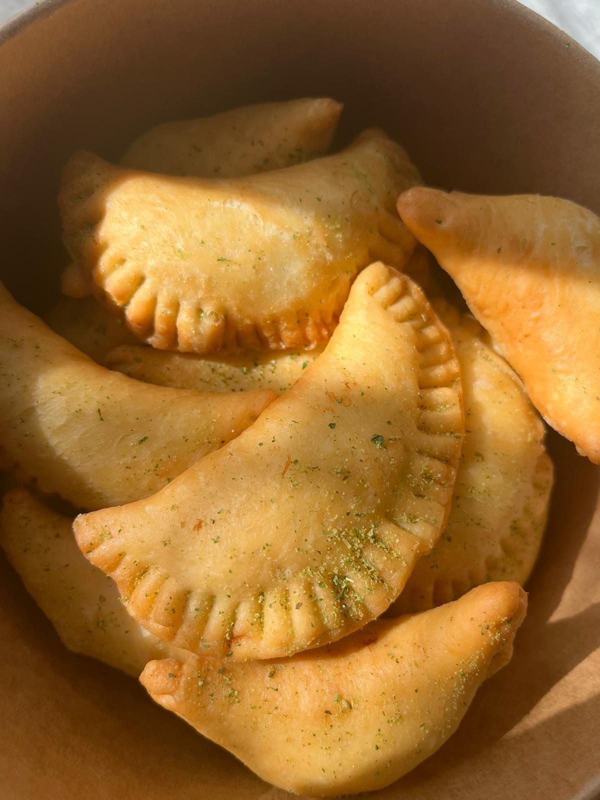 Pâtisseries dorées aux bords ondulés, empilées dans un récipient brun, saupoudrées d'assaisonnement vert.