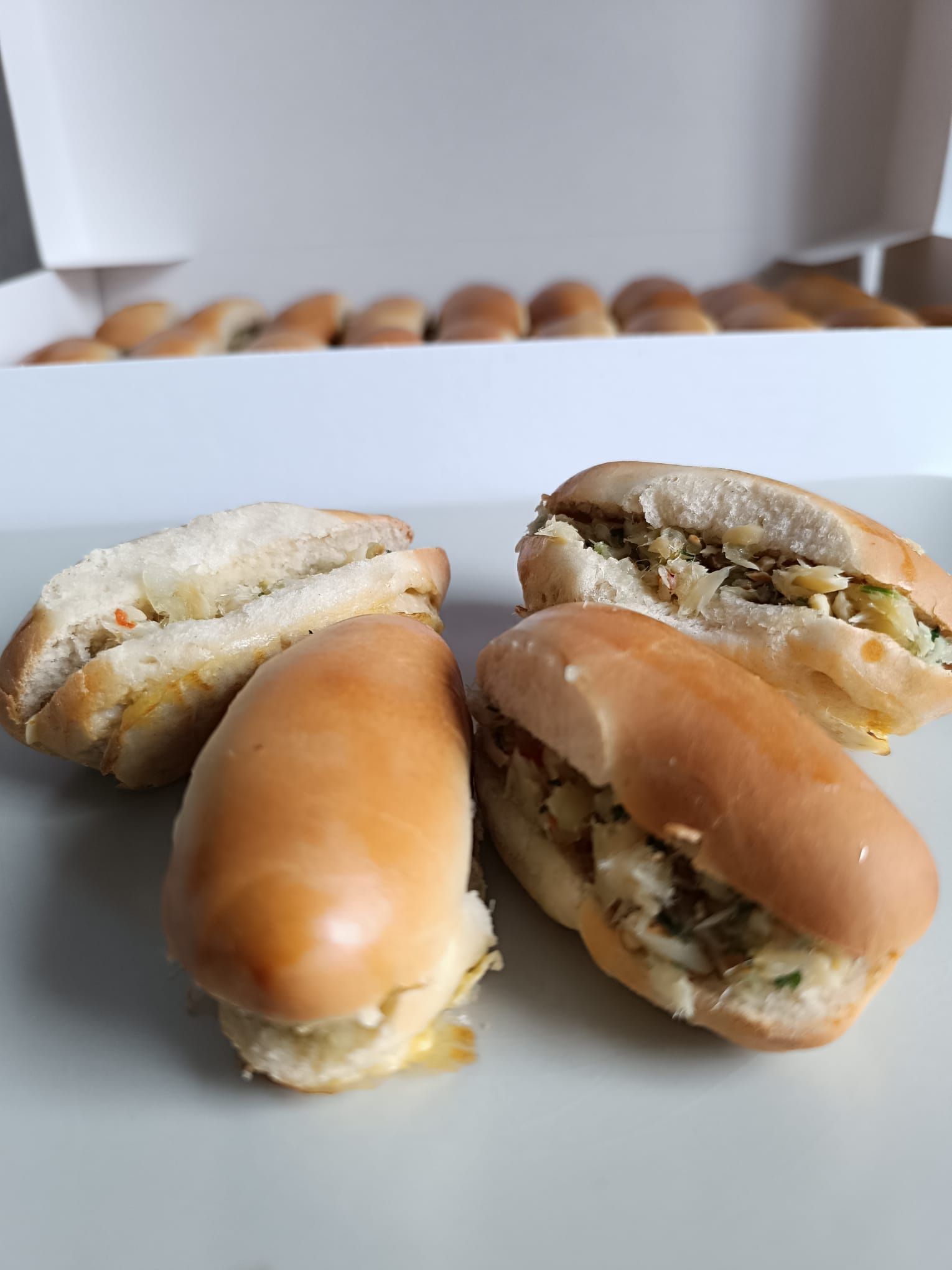 Quatre mini sandwichs sur une assiette blanche, avec d'autres sandwichs dans une boîte blanche en arrière-plan.