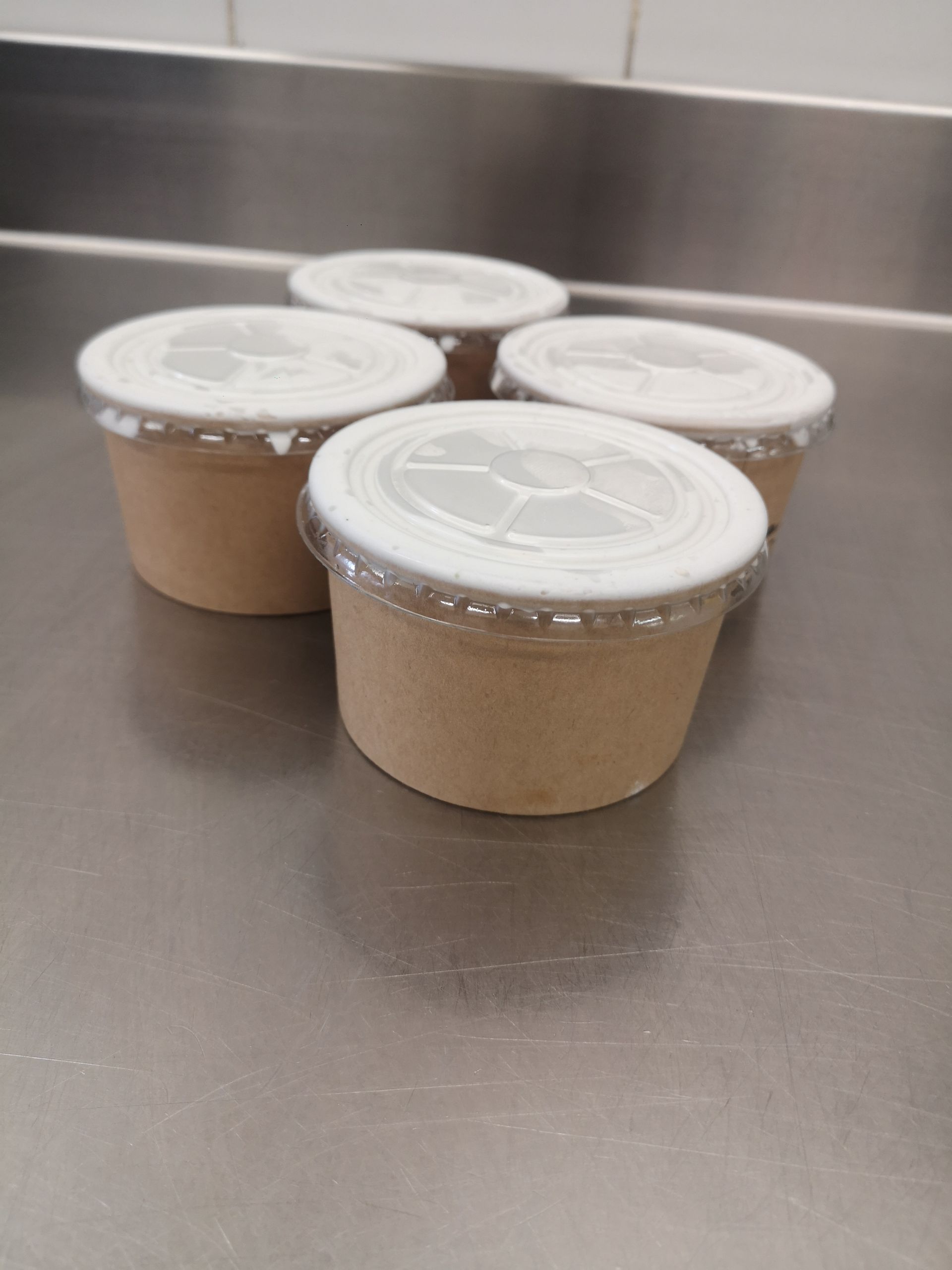 Quatre petits récipients avec couvercle contenant des aliments brun clair sont posés sur une surface en acier inoxydable.