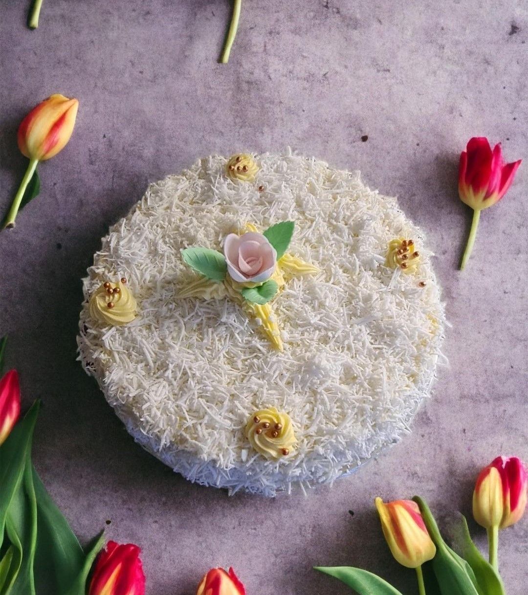 Gâteau avec glaçage blanc, décoré d'une fleur et entouré de tulipes colorées.
