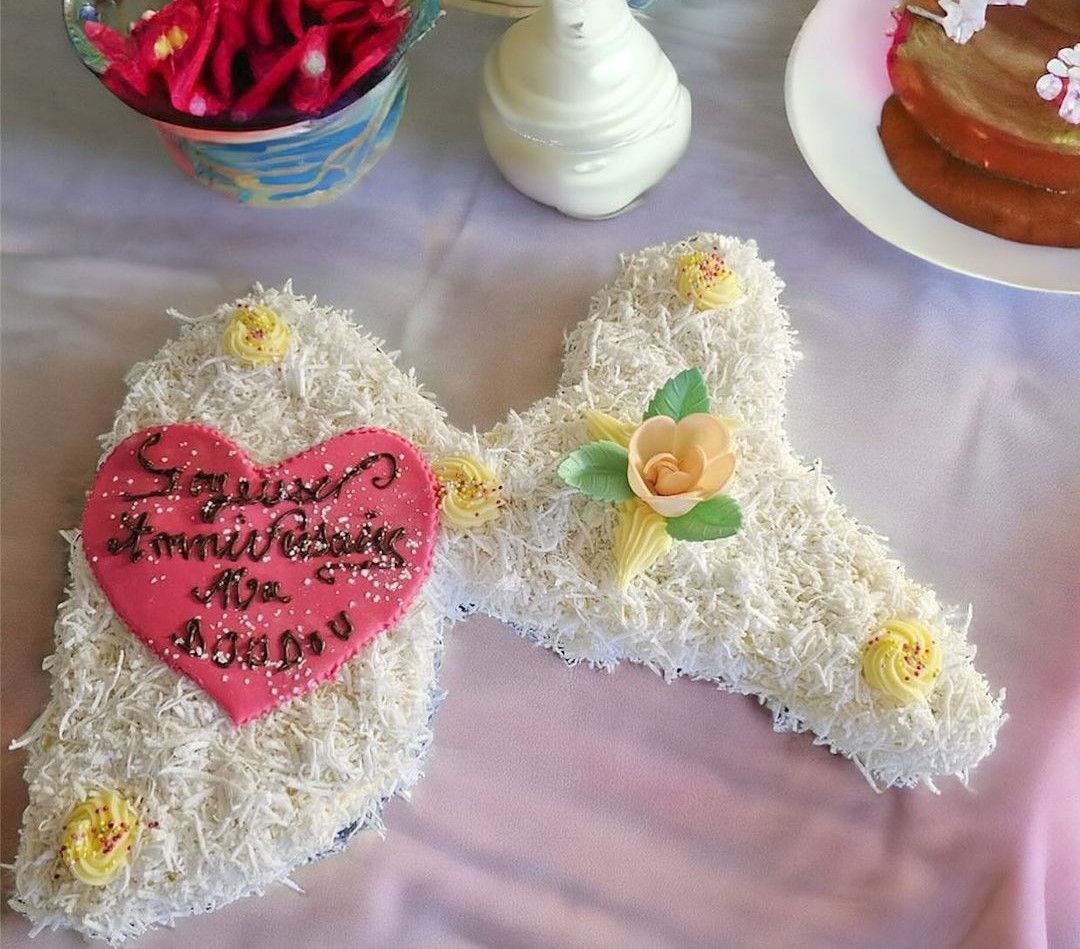 Un gâteau en forme de lettre « H », recouvert de flocons de noix de coco, avec un cœur rose et des décorations de fleurs jaunes.
