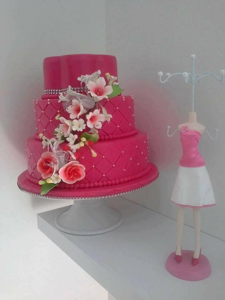 Gâteau rose à trois étages avec des décorations florales sur un support blanc, à côté d'un porte-bijoux en forme de robe.