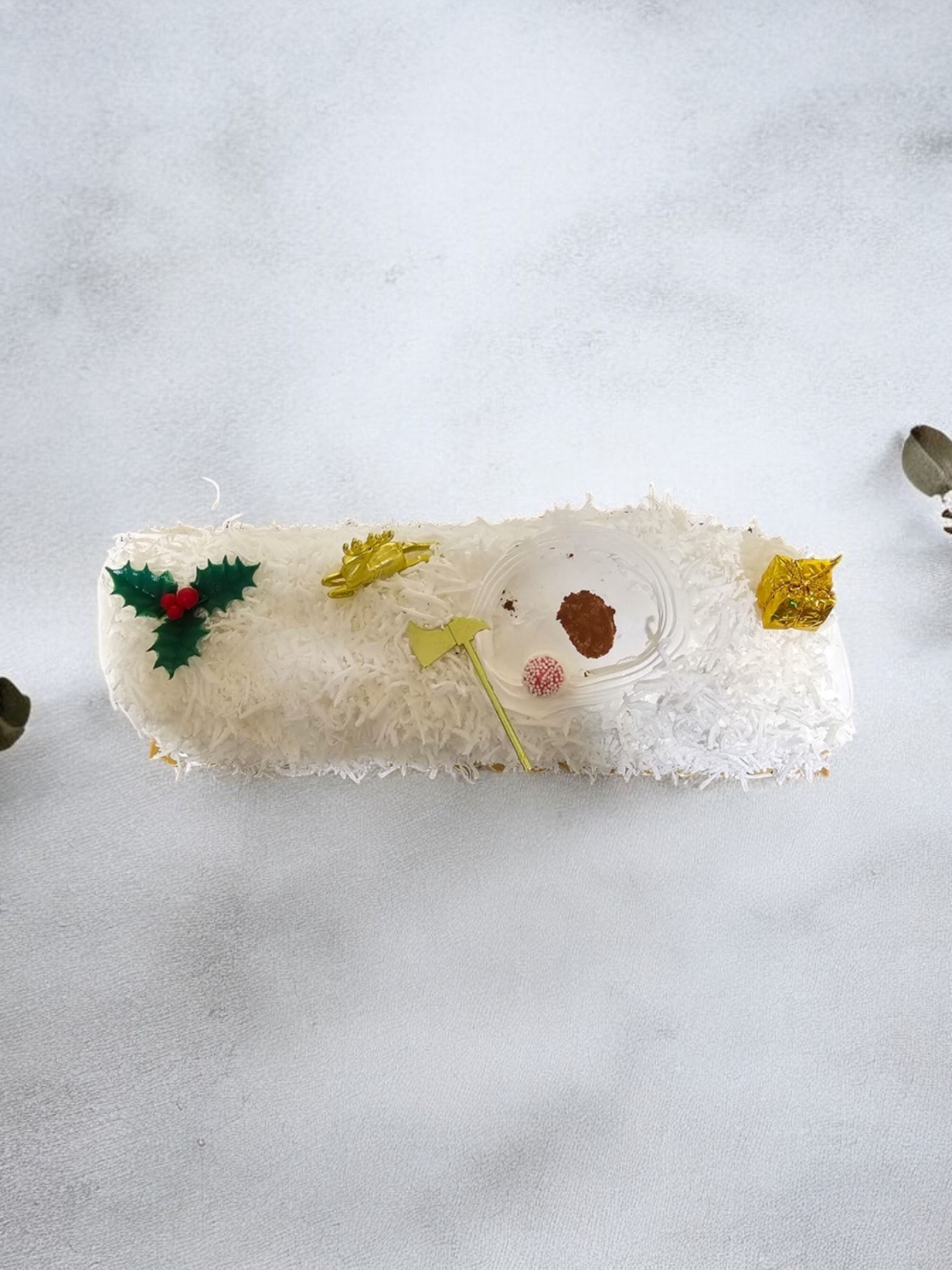 Bûche de Noël blanche, glacée et décorée de houx et de paillettes dorées sur une surface blanche.