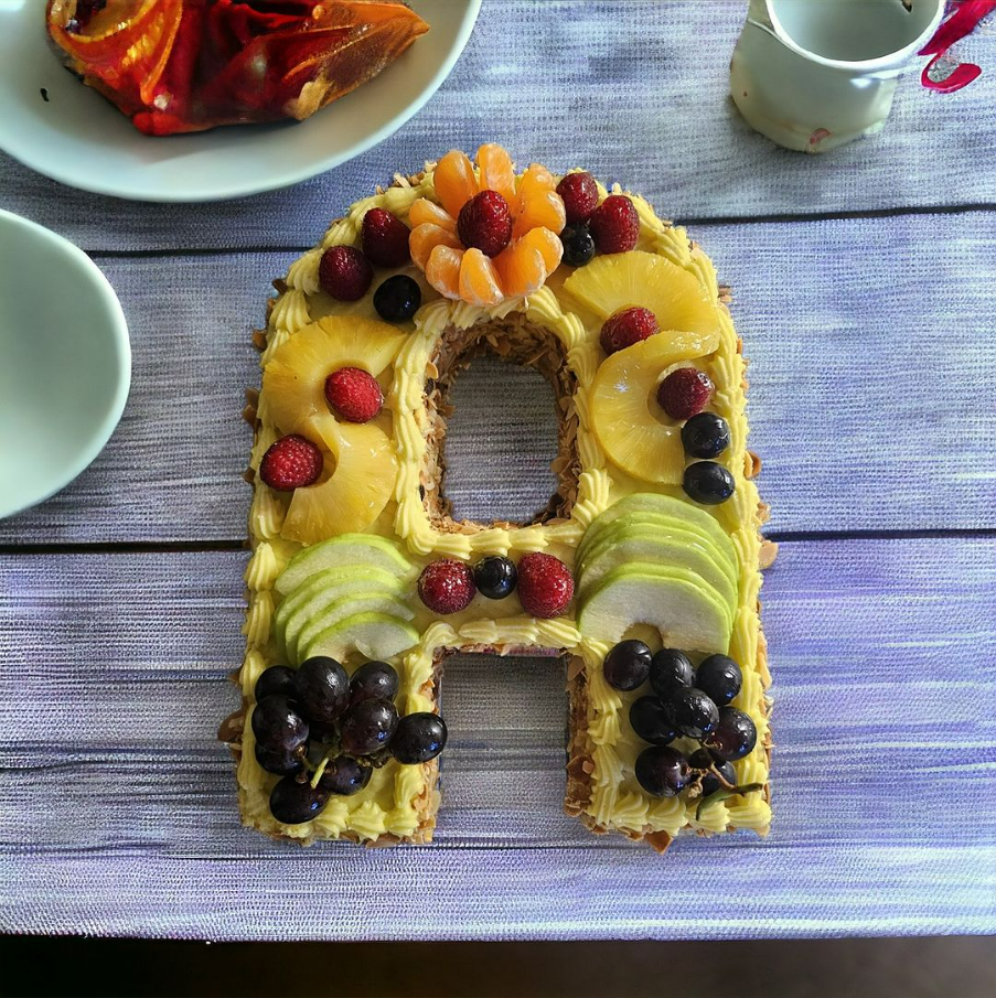 Gâteau en forme de lettre décoré de fruits et de crème sur une table violette, à côté d'assiettes et d'un petit pichet.