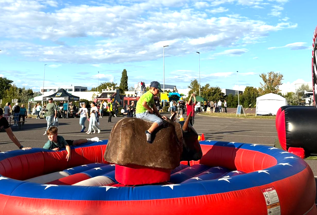 Kinder spielen bei einer Veranstaltung im Freien ein Spiel mit grünen Bechern, im Hintergrund Menschen und eine Bühne.