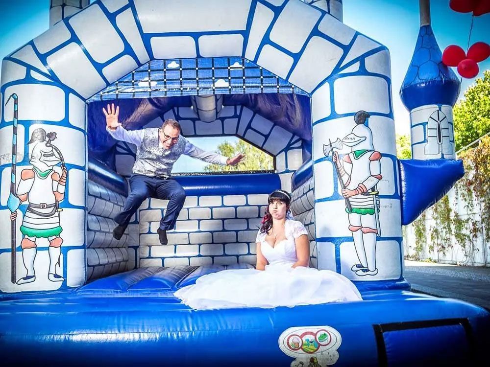 Braut im weißen Kleid sitzt in Hüpfburg, Bräutigam springt. Cartoon-Ritter an den Wänden, Luftballons im Hintergrund.