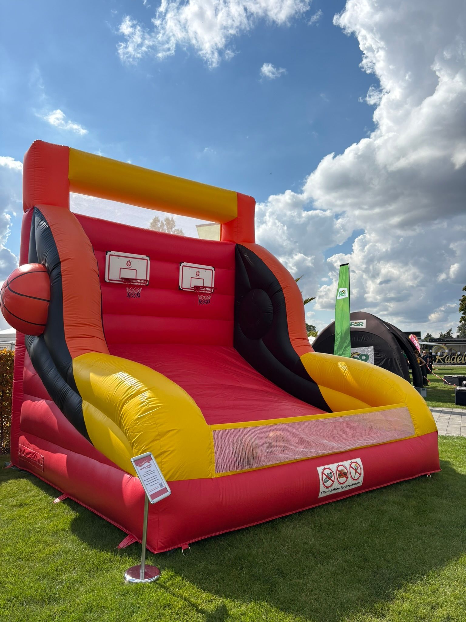 Aufblasbares Basketballspiel mit roten, gelben und schwarzen Feldern, im Freien auf Gras unter blauem Himmel aufgestellt.