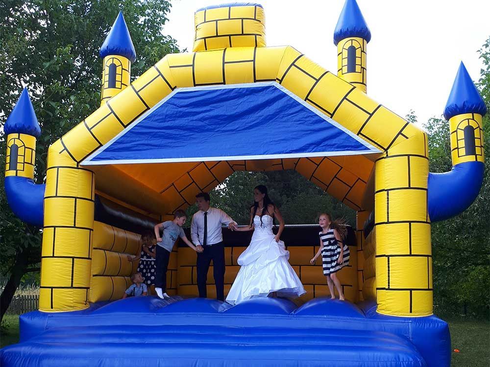 Braut und Bräutigam mit Kindern in einer gelb-blauen Hüpfburg bei einer Hochzeit im Freien.