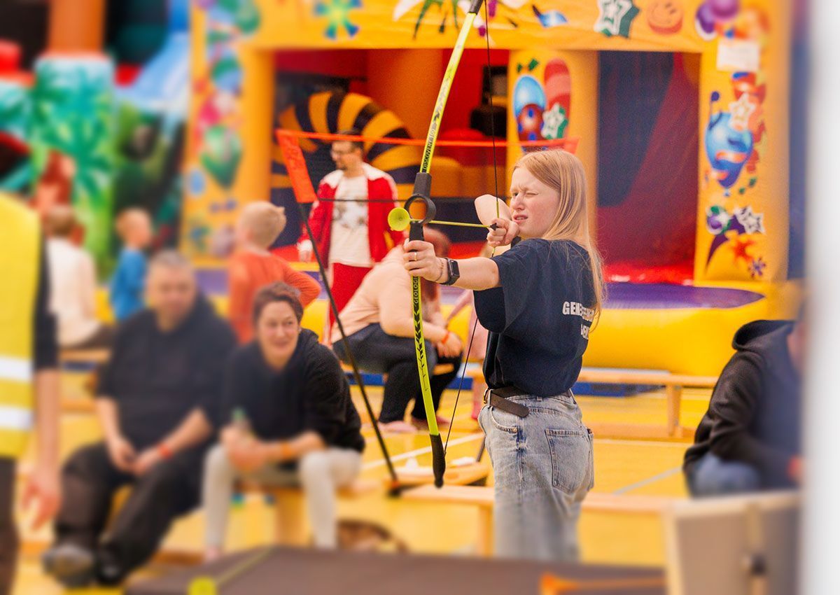 Blondes Mädchen zielt mit Pfeil und Bogen auf eine Zielscheibe in einem Innenbereich. Im Hintergrund sind andere Personen in einem Vergnügungspark zu sehen.