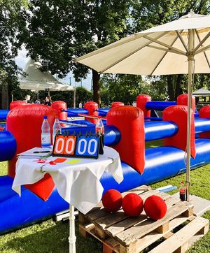 Aufblasbares Tischfußballspiel in einem Park. Anzeigetafel bei 0:0. Rote Bälle, rot-blaue aufblasbare Struktur, kleiner Tisch mit Sonnenschirm.