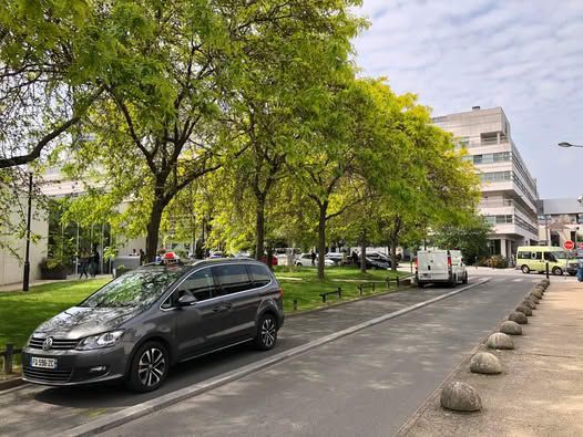 Voiture grise garée sur une route goudronnée bordée d'arbres, avec un bâtiment en arrière-plan.