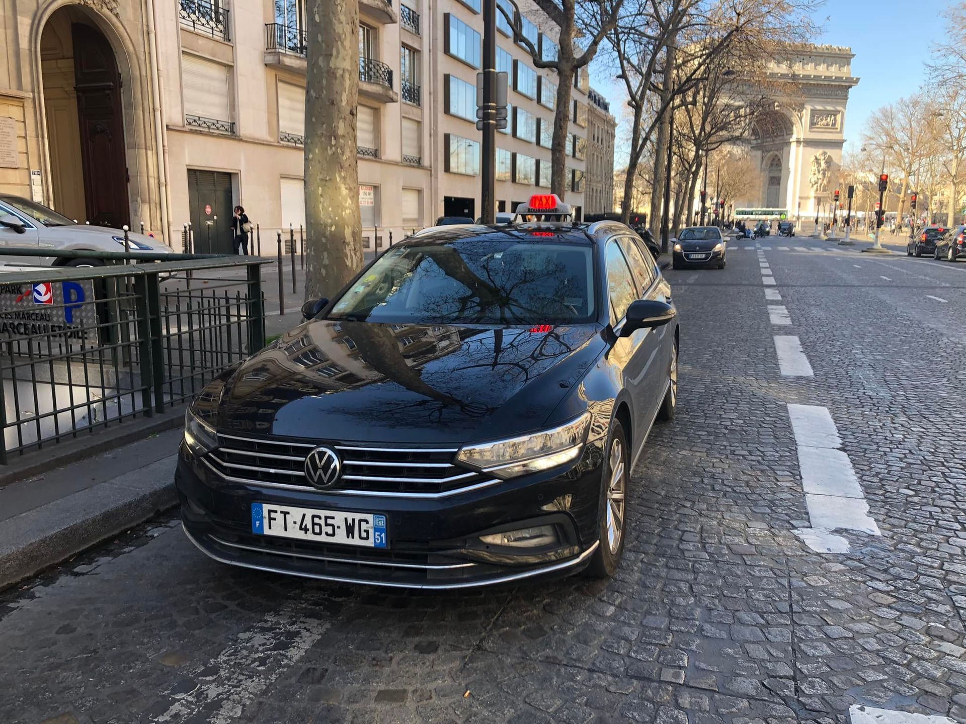 Une berline noire est garée dans une rue pavée de Paris, avec l'Arc de Triomphe en arrière-plan.