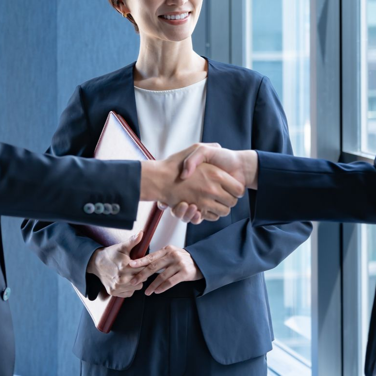Deux personnes se serrent la main, une femme en tailleur sourit et tient un classeur près d'une fenêtre.
