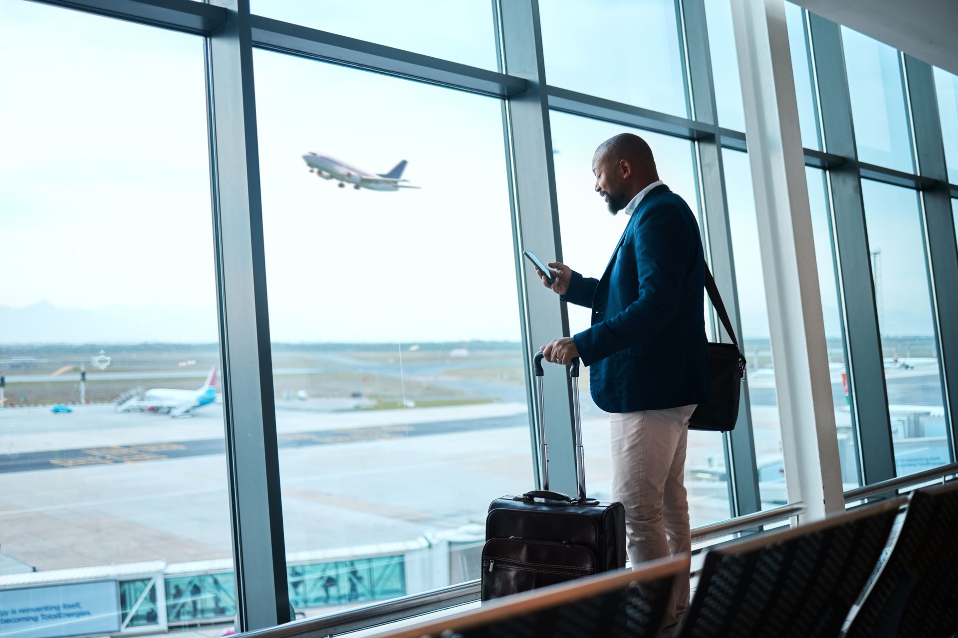 Un homme en costume, devant la fenêtre d'un aéroport, regarde un avion décoller.