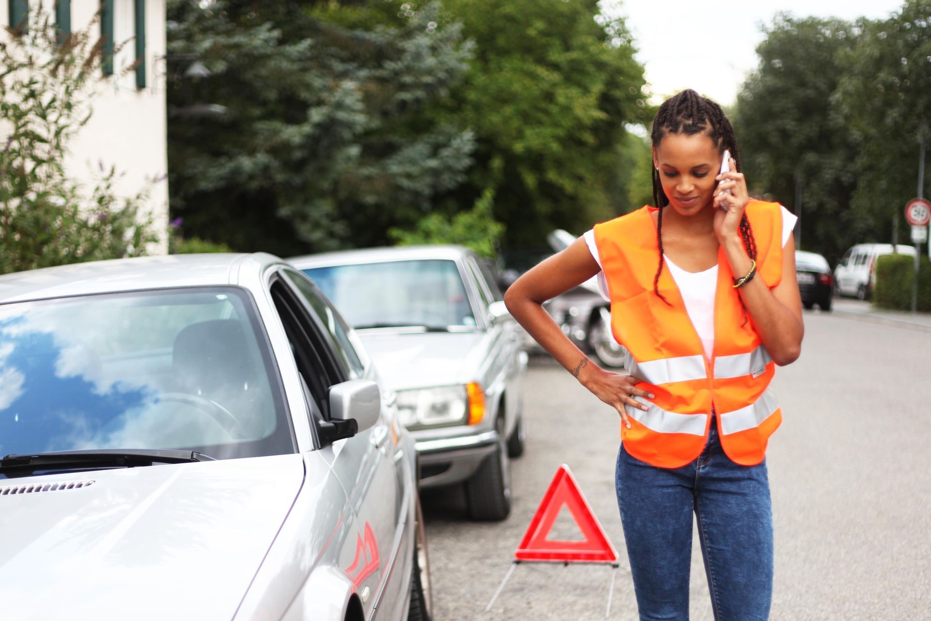 Une femme en gilet orange, au téléphone, près d'une voiture avec un triangle de signalisation sur la chaussée.