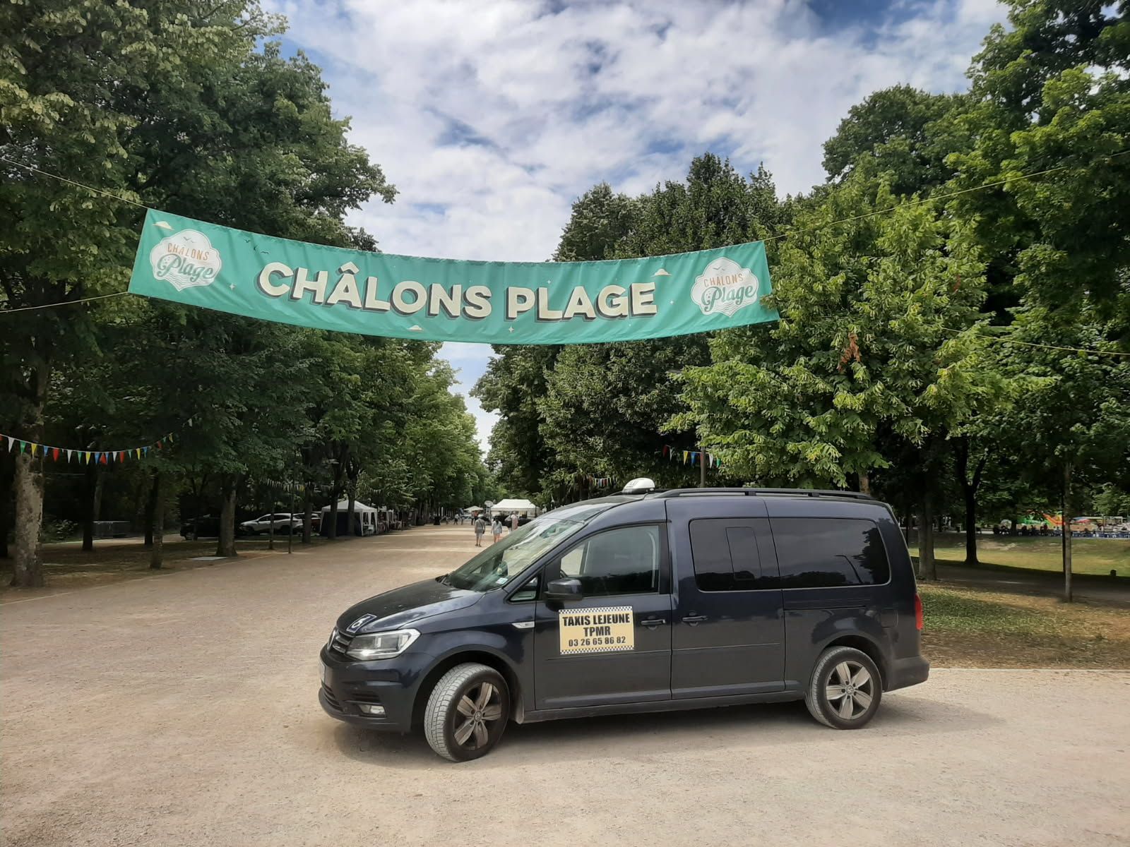 Un taxi van sombre est garé sur un chemin de gravier sous une banderole indiquant Châlons Plage.