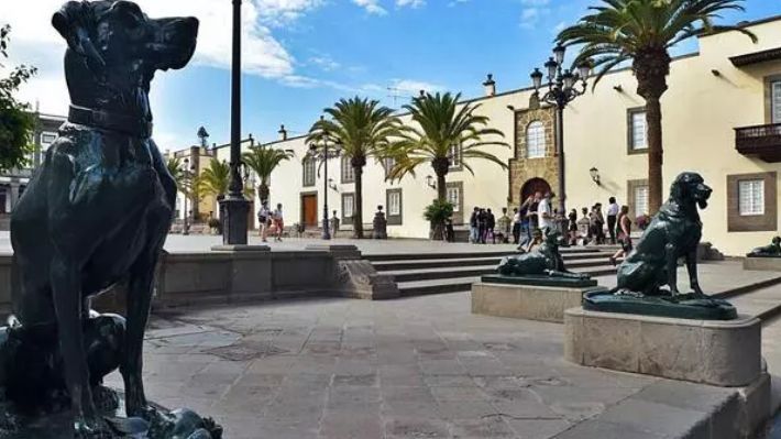 Estatuas de perros de bronce sobre pedestales en una plaza soleada con palmeras y edificios de estilo colonial de color crema.
