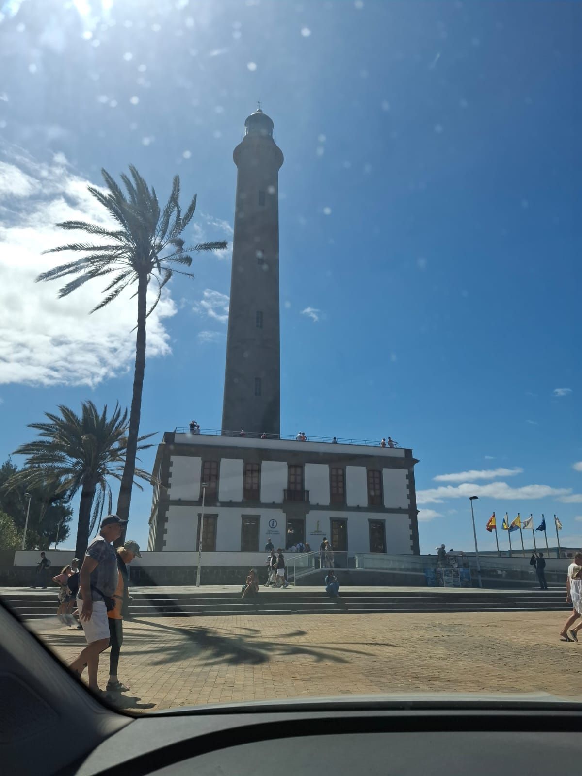 Un faro alto y cilíndrico con una base blanca se alza bajo un cielo azul despejado, junto a una palmera en una plaza pavimentada.