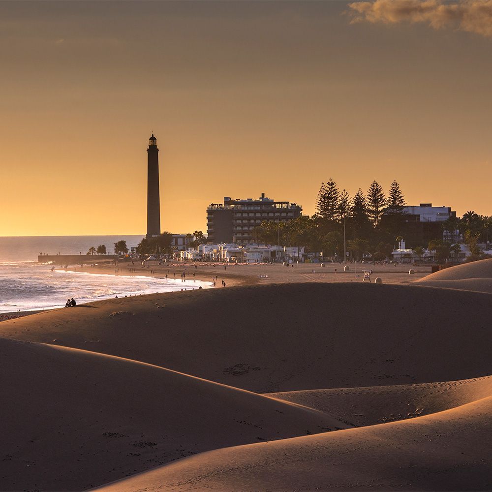Puesta de sol dorada sobre las dunas de Maspalomas, en las Islas Canarias, con el emblemático faro visible a lo lejos.