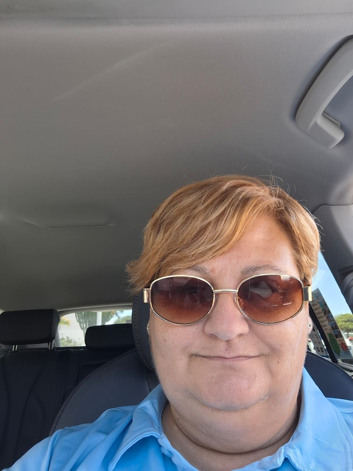 Una persona con cabello corto y castaño claro, que lleva gafas de sol con cristales marrones y una camisa azul con cuello, se encuentra dentro de un automóvil.