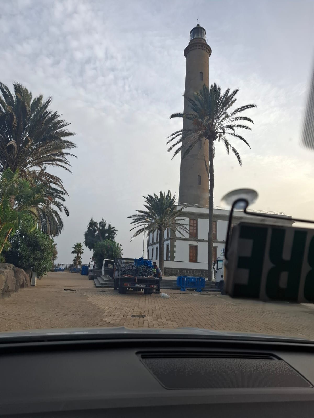 Una alta torre de faro de color claro se alza cerca de palmeras en una zona costera, vista desde el interior de un coche.