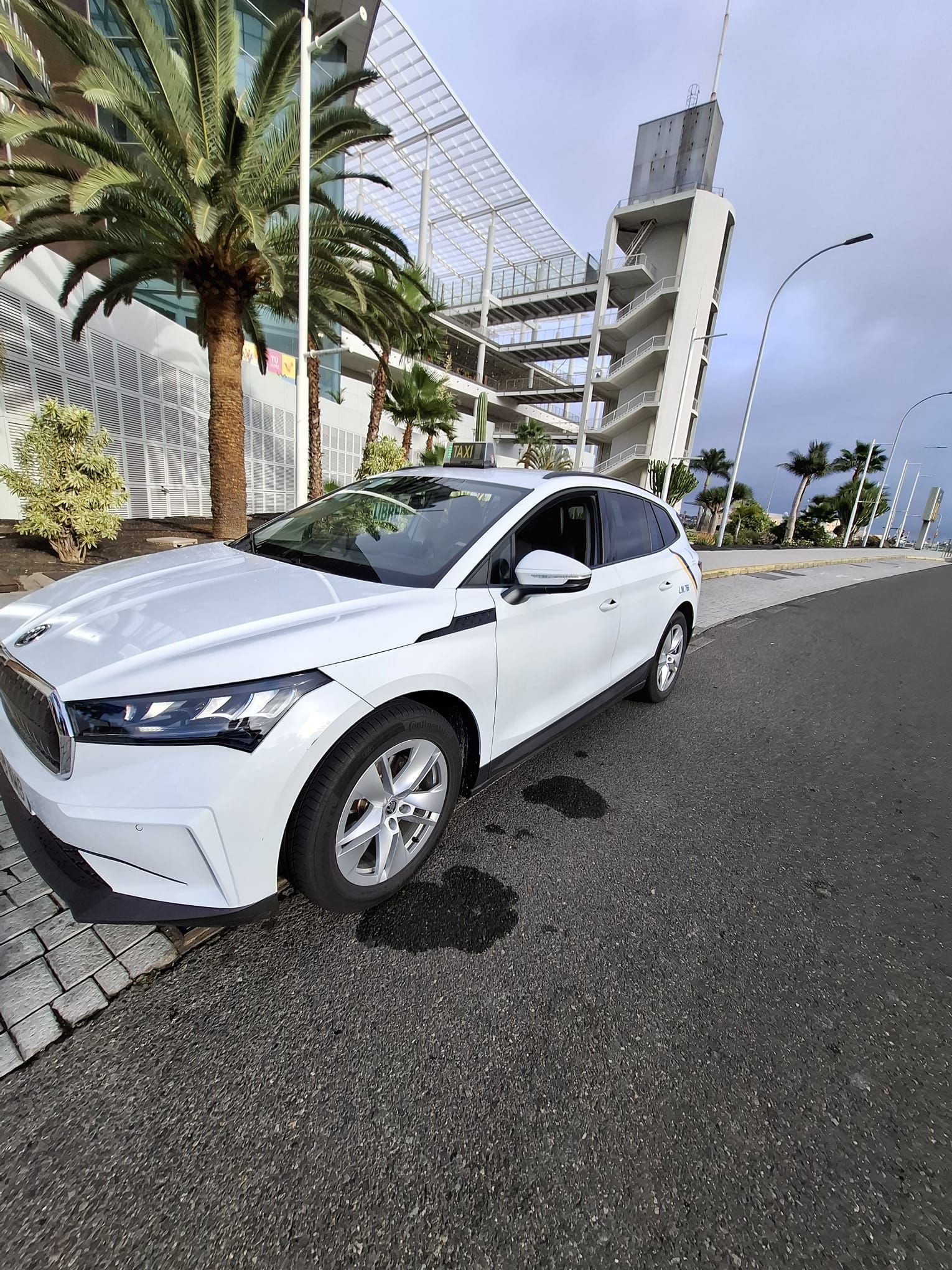 Un SUV blanco está estacionado al costado de una carretera cerca de un gran estadio moderno con palmeras bajo un cielo nublado.