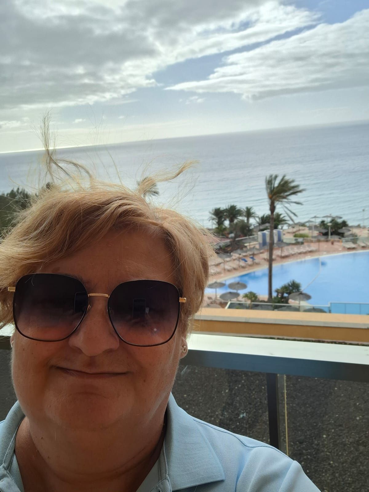 Una persona con gafas de sol sonríe frente a la barandilla de un balcón con vistas a una piscina y al océano.