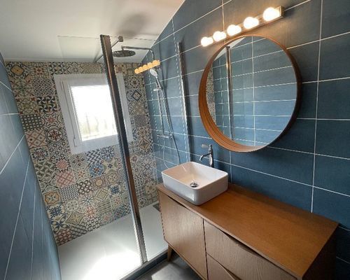 salle de bain carrelage bleu moderne, meuble bois avec vasque a poser blanche à droite,  parois de douche au fond miroir rond éclairer a droite.