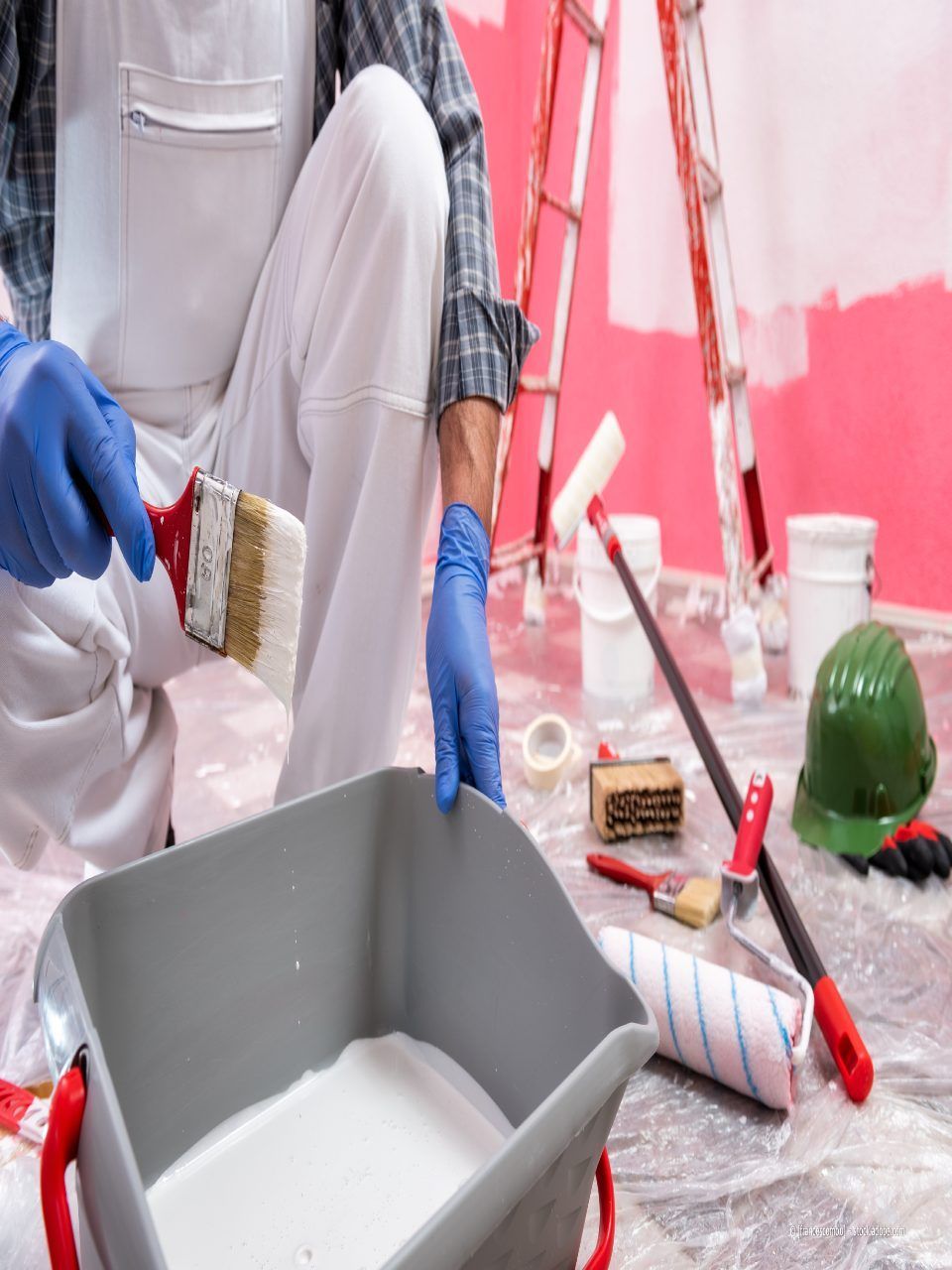 Eine Person, die mit Pinsel und blauen Handschuhen eine Wand streicht, ist umgeben von Malutensilien und einem grauen Tablett.