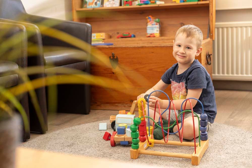 Ein Junge spielt auf einem Teppich mit einem Labyrinth aus Holzperlen und Bauklötzen; im Hintergrund sind eine Holztruhe und ein Regal zu sehen.