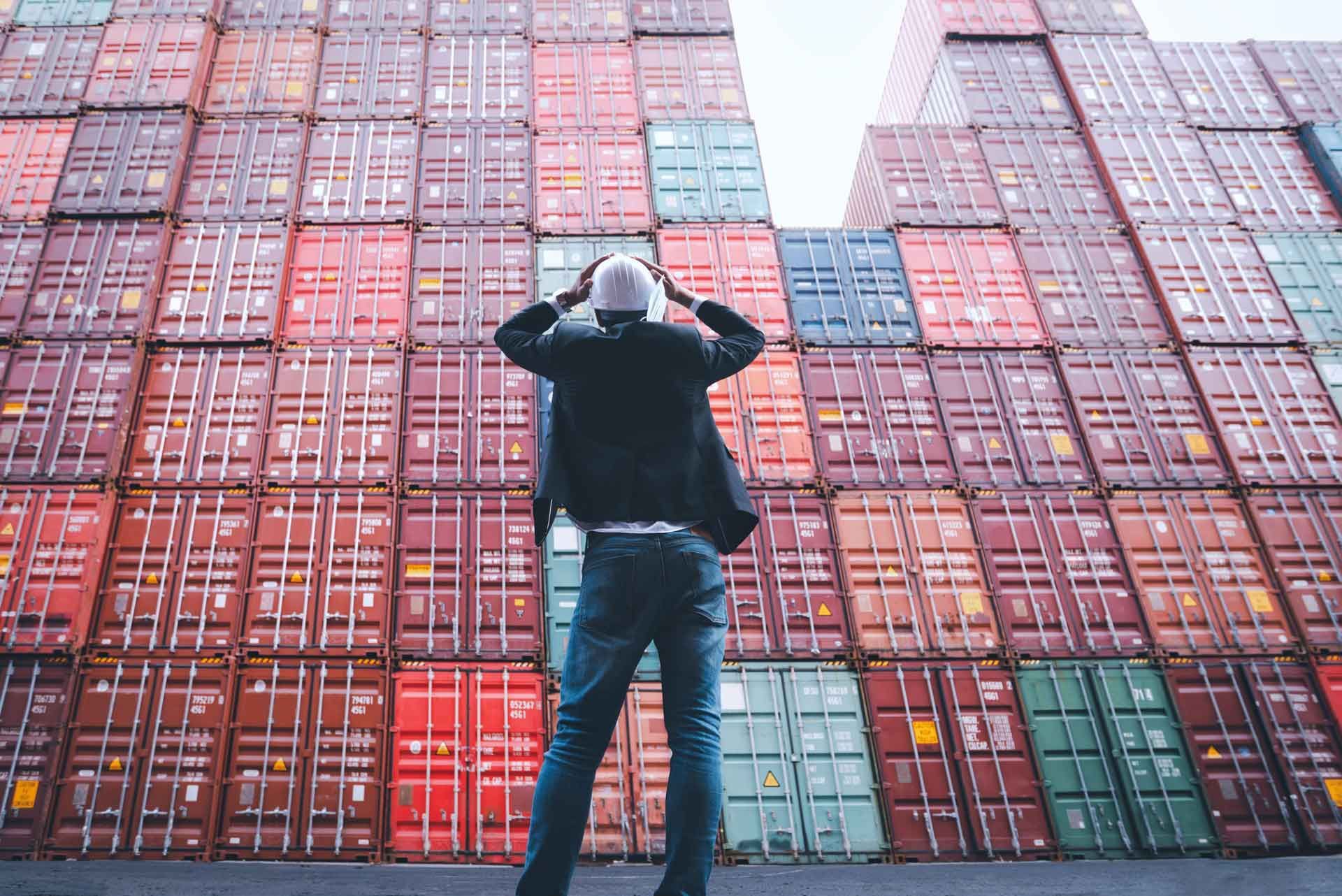 Un homme vu de dos se tenant la tête devant un amoncellement de conteneurs maritimes