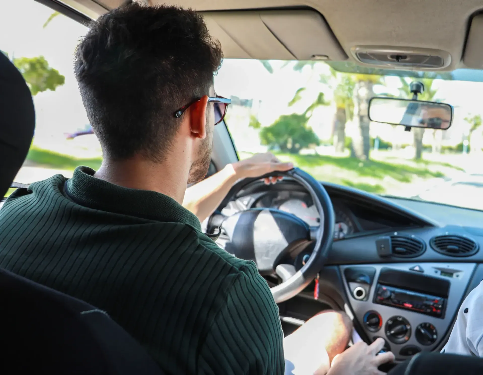 Persona conduciendo un coche, con las manos en el volante, vista desde el asiento trasero. Luz diurna, techo verde.
