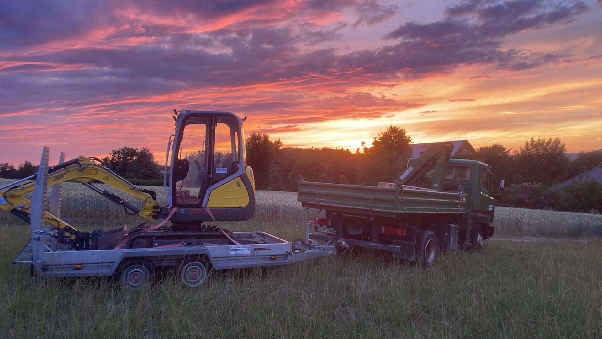 Ein gelber Bagger auf einem Anhänger und ein grüner Muldenkipper parken bei Sonnenuntergang auf einer Wiese.