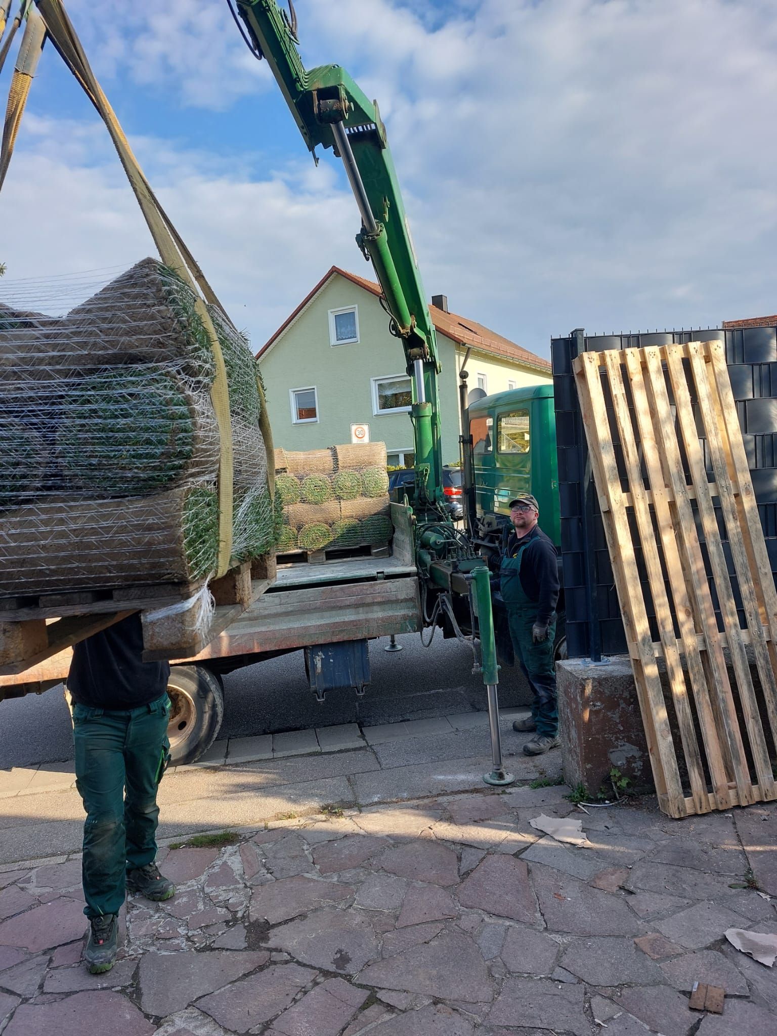 Arbeiter laden mit einem Kran vor einem Gebäude Rasensoden von einem LKW ab.