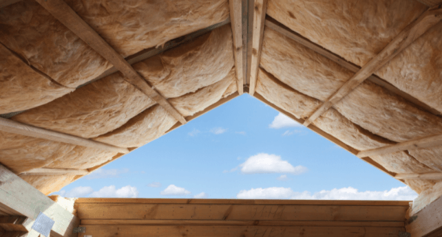 Vue intérieure d'un grenier non aménagé avec poutres en bois apparentes, isolation et une ouverture triangulaire donnant sur le ciel bleu.