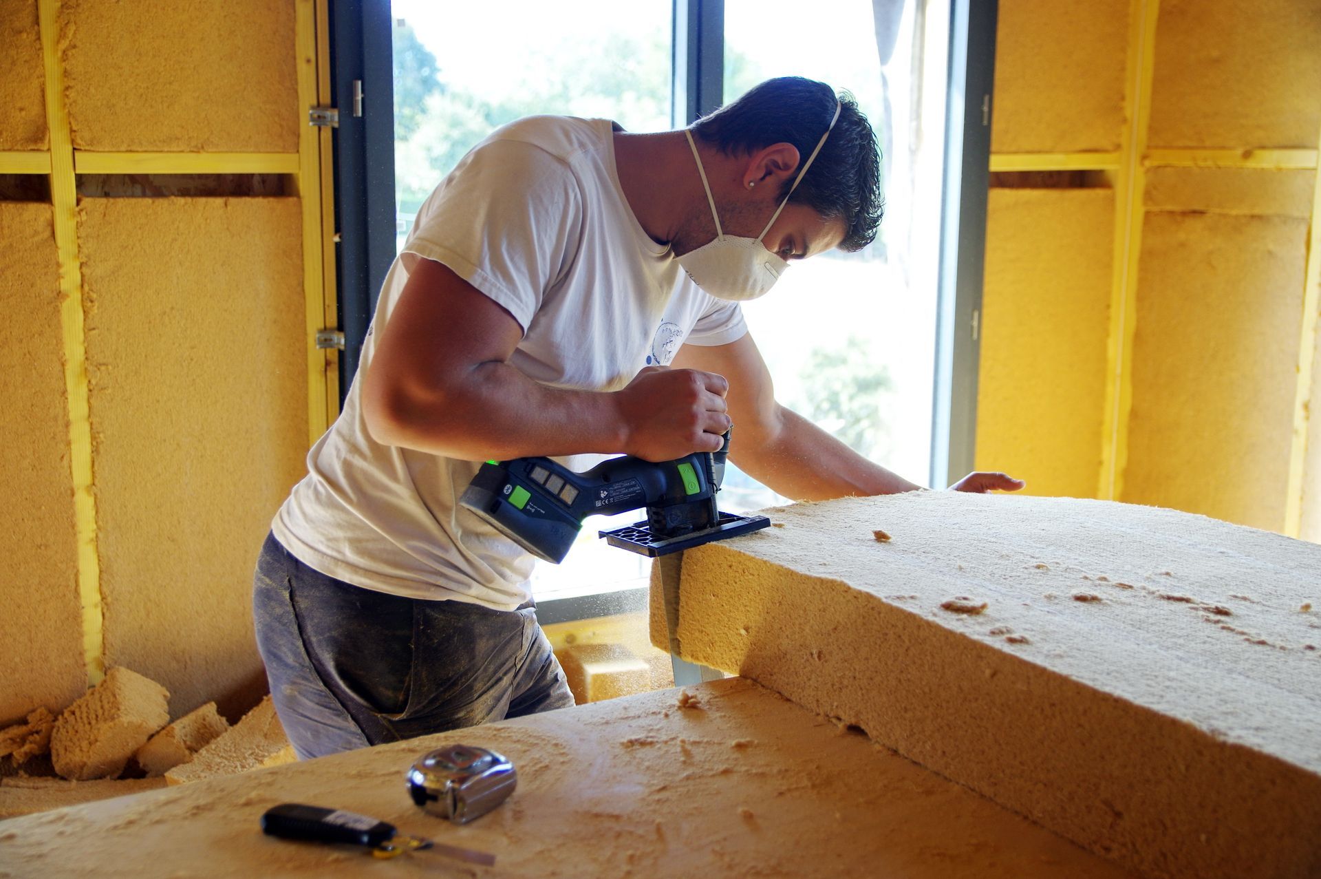 Un homme portant un masque utilise une scie sur un matériau isolant à l'intérieur d'un bâtiment comportant une fenêtre.