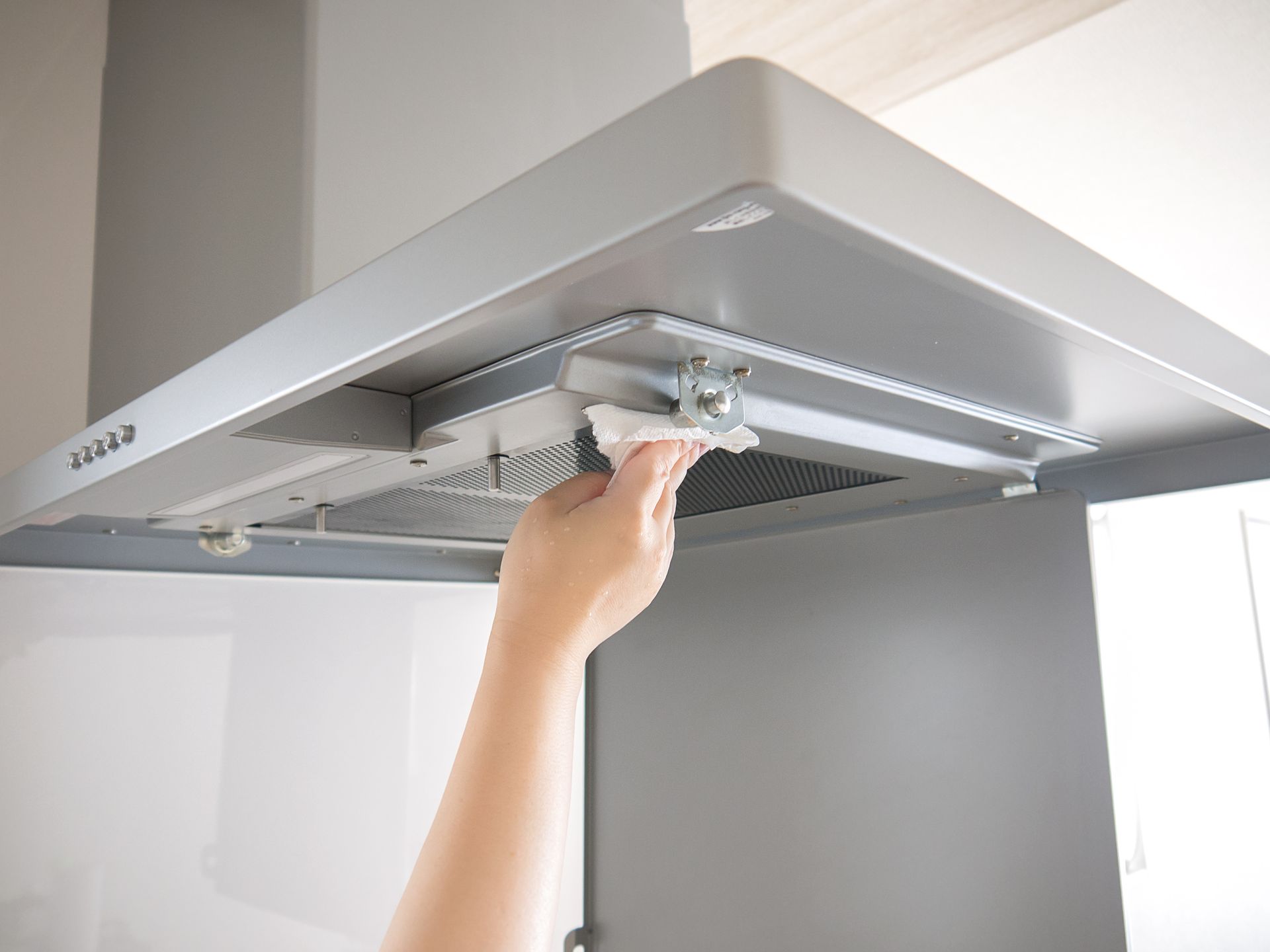 Personne nettoyant une hotte de cuisinière argentée dans une cuisine blanche avec un chiffon.