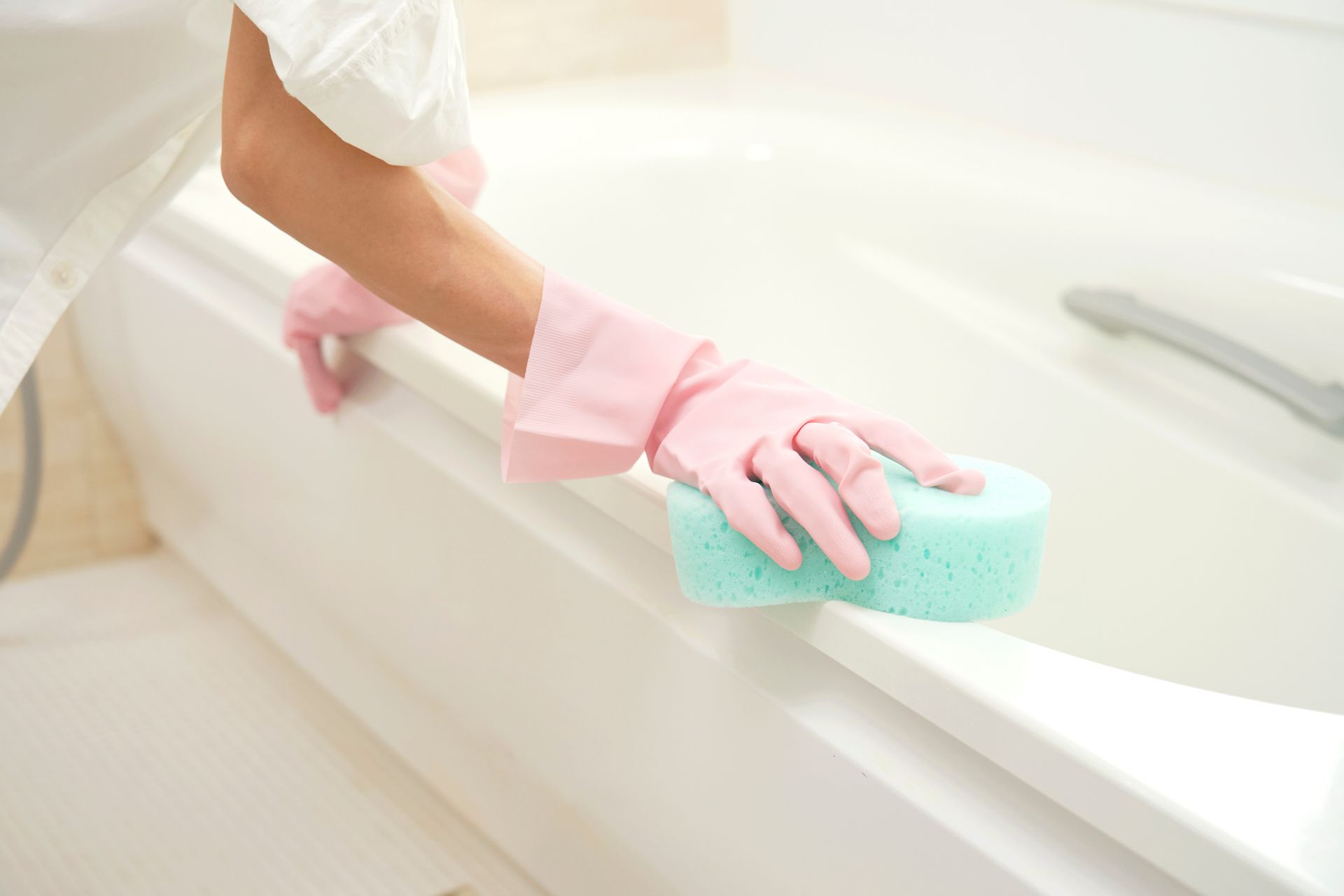 Une personne portant des gants roses frotte le bord d'une baignoire blanche avec une éponge bleue.