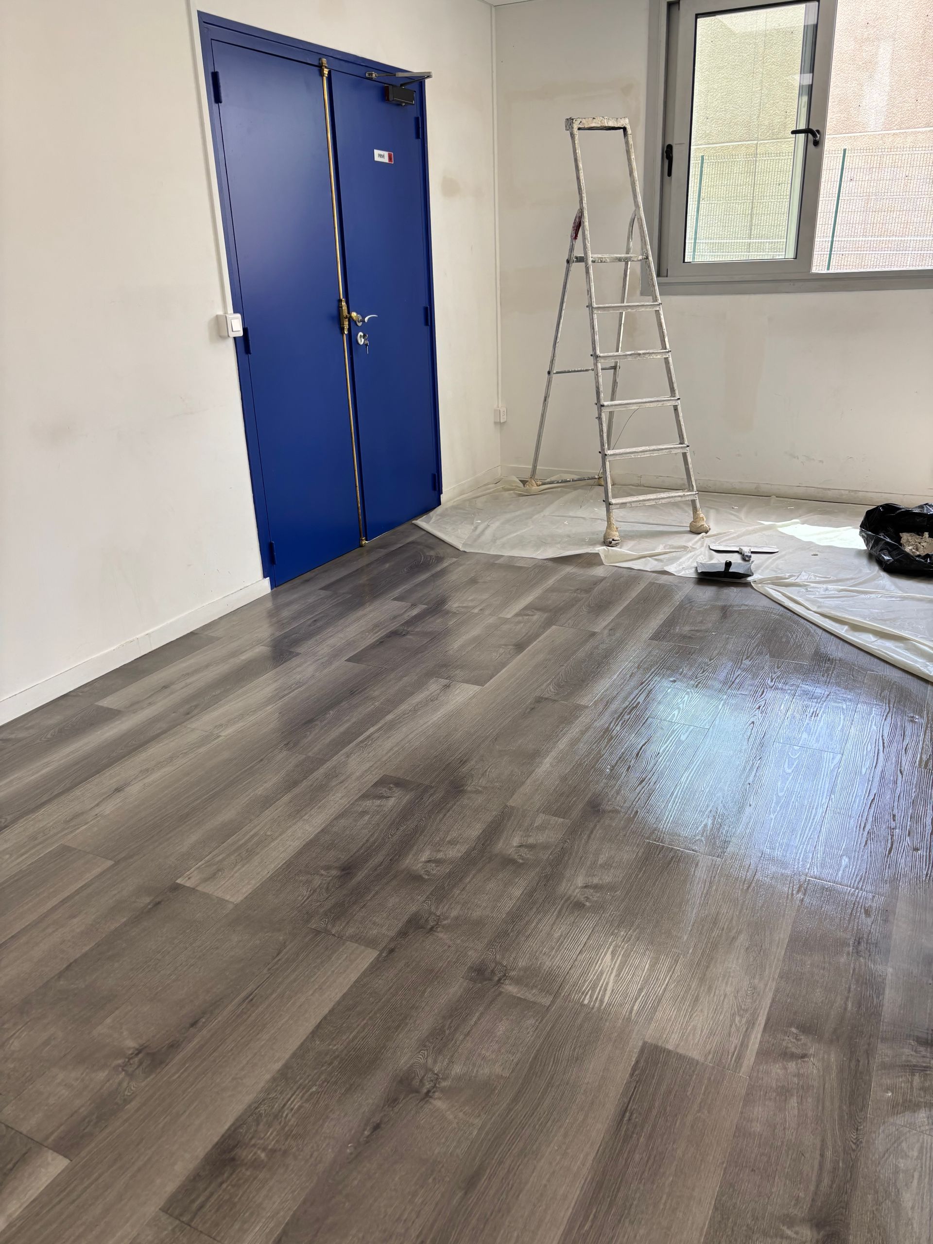 Une personne passe l'aspirateur sur le sol poussiéreux d'une pièce, probablement en construction, à l'aide d'un tuyau relié à un aspirateur visible.