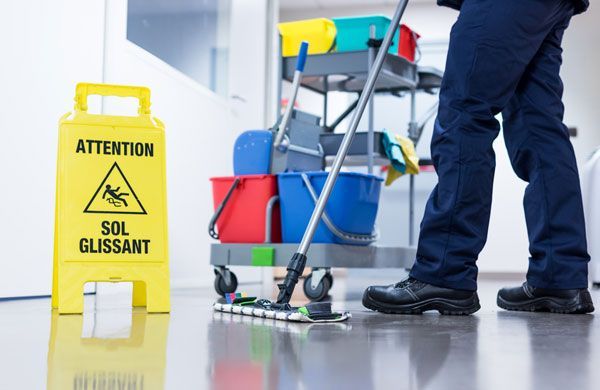 Une personne nettoie un sol brillant à côté d'un panneau indiquant un sol mouillé et d'un chariot de nettoyage.