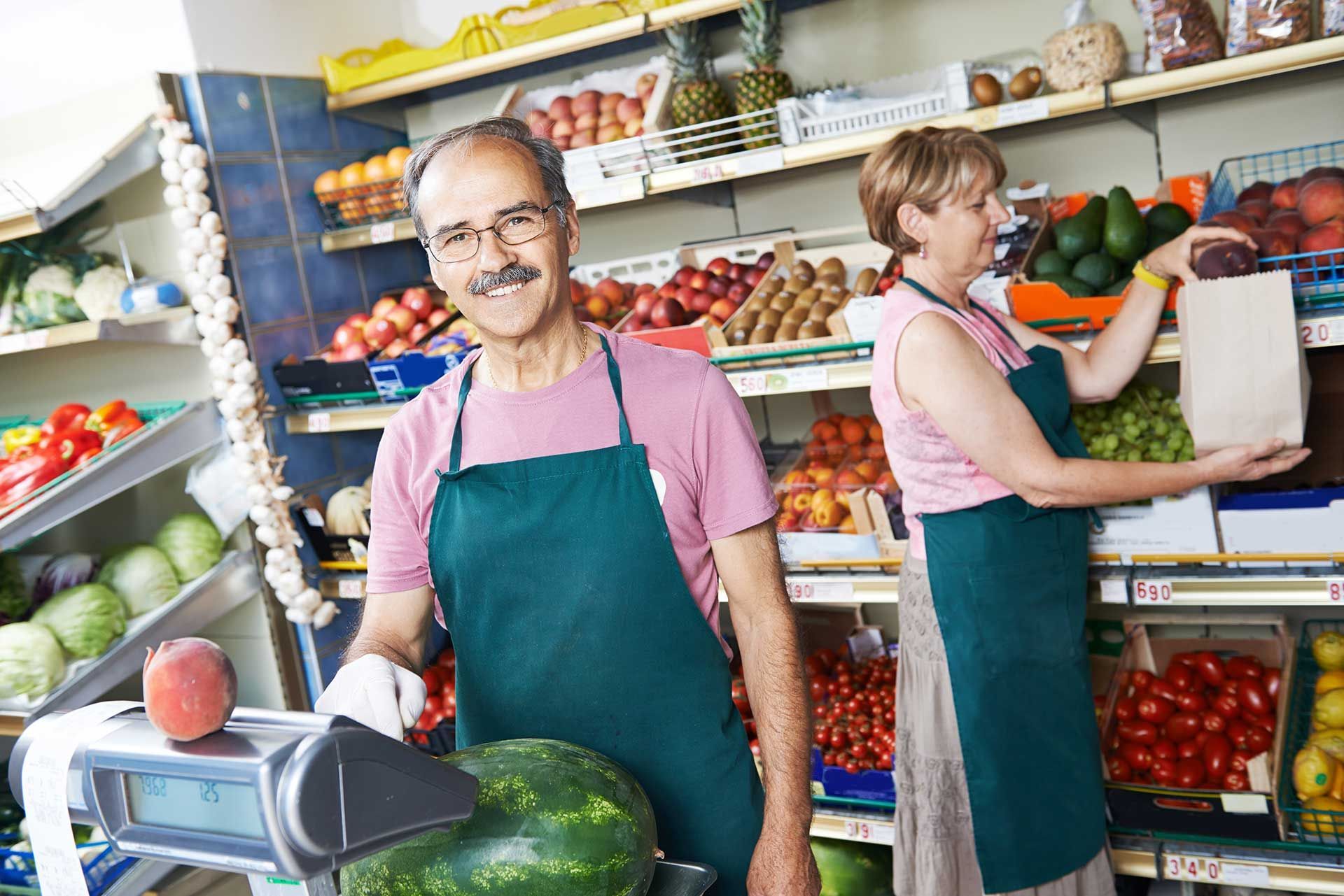 Deux personnes en tablier vert devant un étal de fruits. L'une pèse une pastèque, l'autre ensache les fruits et légumes.