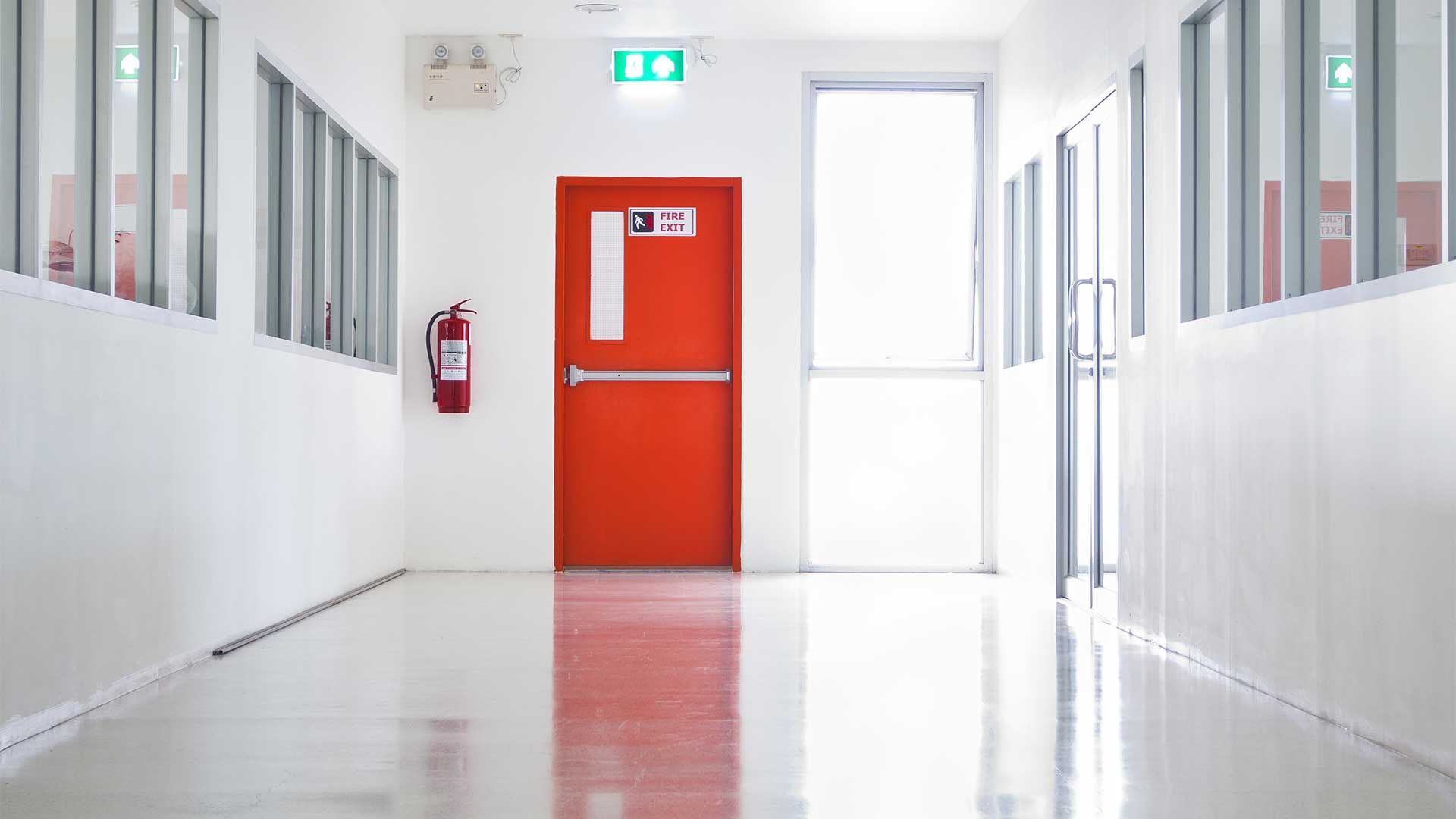 Long couloir avec porte coupe-feu rouge, extincteur et panneau de sortie.