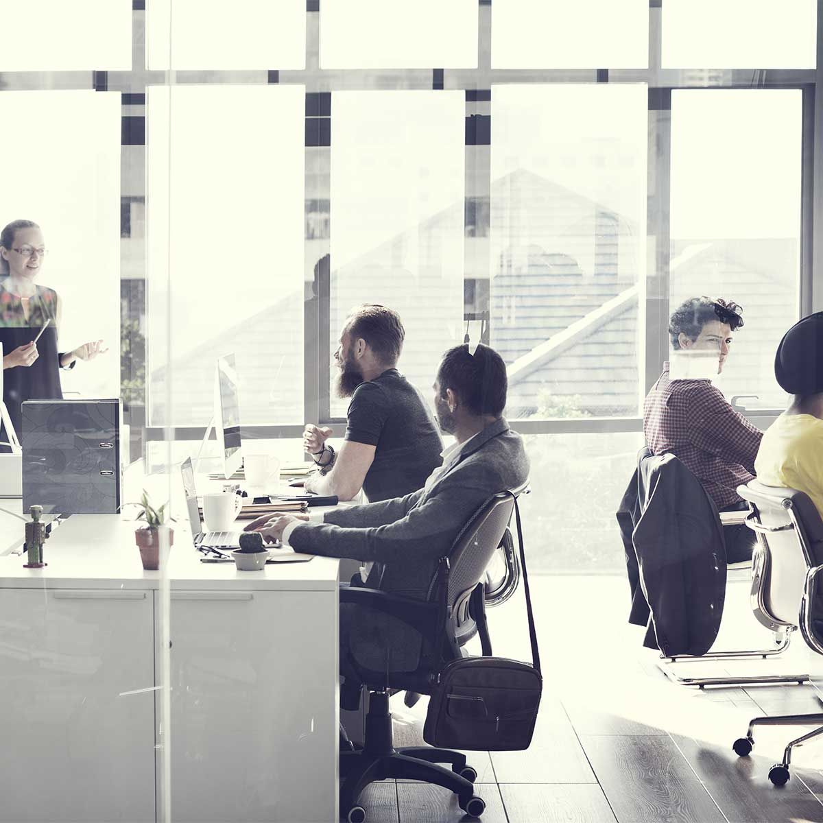 Bureau avec des employés travaillant à des bureaux près d'une fenêtre. Une femme se tient à l'arrière-plan et fait une présentation.