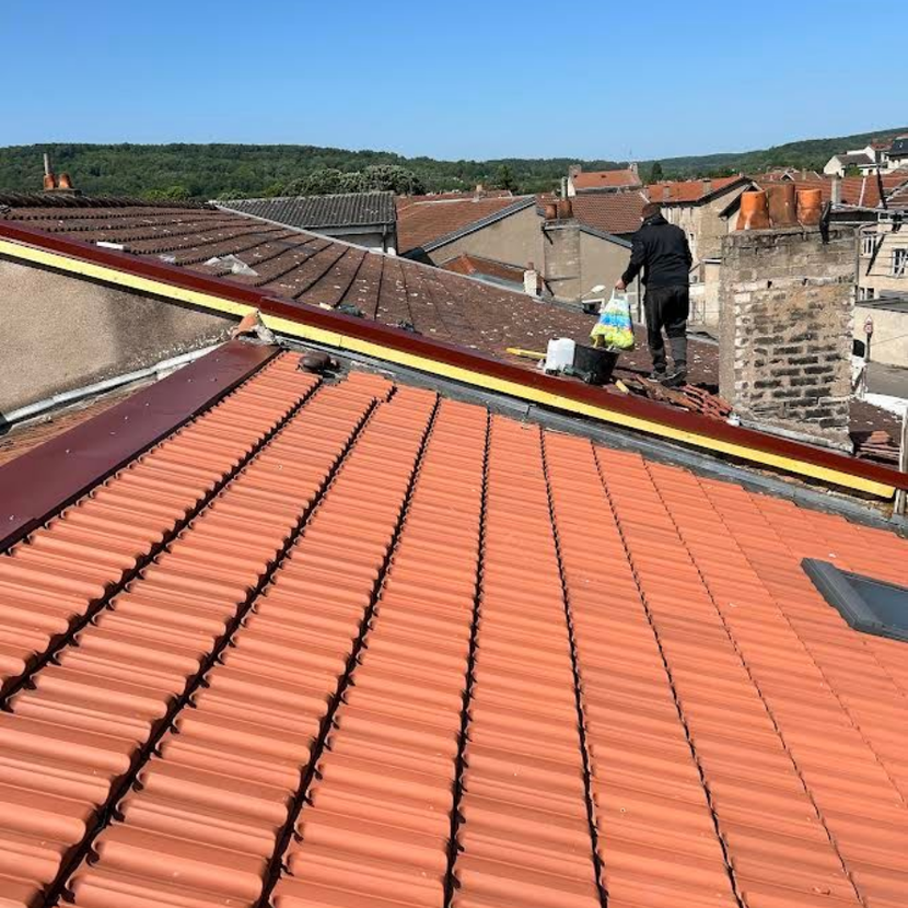 Un homme répare une partie d'un toit de tuiles en terre cuite, avec des toits et des arbres en arrière-plan.
