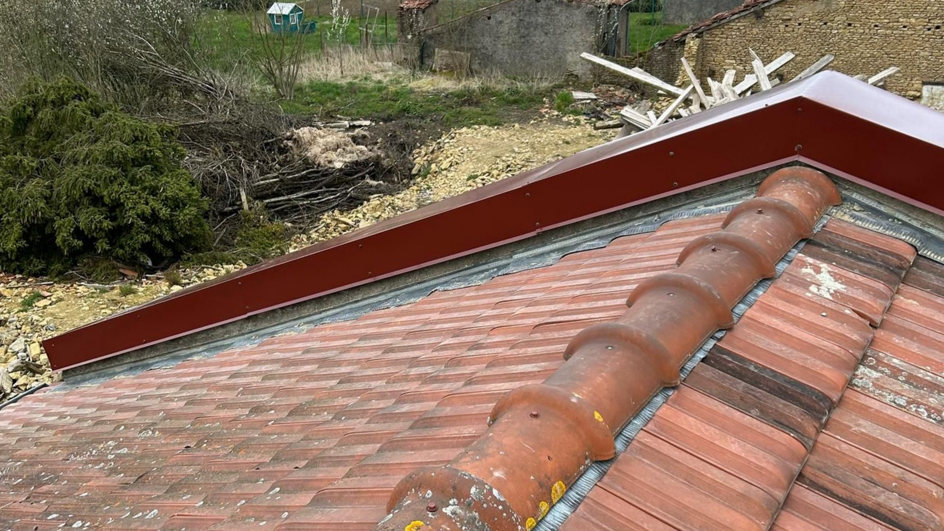 Toit en tuiles rouges avec une corniche en métal rouge ; arrière-plan de végétation envahissante et de matériaux de construction.